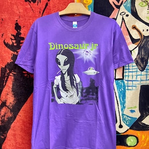 May include: A purple t-shirt featuring the band name "Dinosaur Jr" in green text. The graphic depicts an alien smoking a cigarette, a UFO, and a burst of light. The shirt is hanging on a wooden hanger.