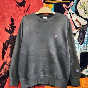 Puede incluir: Sudadera Champion gris oscuro con un pequeño logotipo en el pecho y la manga. La sudadera de cuello redondo está colgada de una percha de madera. El fondo presenta un diseño abstracto colorido.