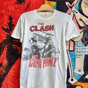 以下が含まれることがあります： クリーム色の半袖Tシャツで、軍服姿の2人の人物が描かれたグラフィックプリントが特徴です。画像の上部には赤色で「THE CLASH」の文字があり、人物の下には赤色で「OUT OF CONTROL」という言葉がプリントされています。