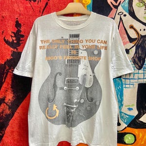 Puede incluir: Camiseta blanca con un gran gr&aacute;fico de guitarra gris y el texto "THE ONLY THING YOU CAN REALLY FEEL IN YOUR LIFE IS NIGO'S FAVORITE SHOP". La camiseta est&aacute; colgada en una percha de madera.