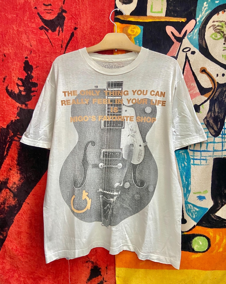 Puede incluir: Camiseta blanca con un gran gr&aacute;fico de guitarra gris y el texto "THE ONLY THING YOU CAN REALLY FEEL IN YOUR LIFE IS NIGO'S FAVORITE SHOP". La camiseta est&aacute; colgada en una percha de madera.