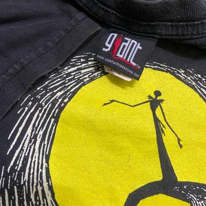 May include: Black t-shirt featuring a graphic of a character standing in front of a yellow moon. The design is in white and black. The tag reads "Giant" and "Large".