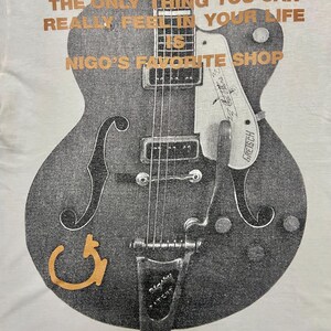 Puede incluir: Camiseta blanca con un gr&aacute;fico de guitarra desgastada en gris y negro. El texto dorado dice: "THE ONLY THING YOU CAN REALLY FEEL IN YOUR LIFE IS NIGO'S FAVORITE SHOP." El dise&ntilde;o de la guitarra incluye la marca "Gretsch".