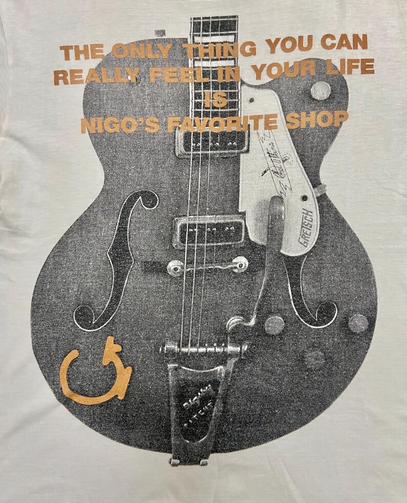Puede incluir: Camiseta blanca con un gr&aacute;fico de guitarra desgastada en gris y negro. El texto dorado dice: "THE ONLY THING YOU CAN REALLY FEEL IN YOUR LIFE IS NIGO'S FAVORITE SHOP." El dise&ntilde;o de la guitarra incluye la marca "Gretsch".
