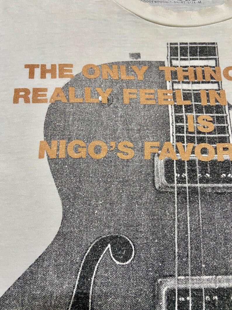 Puede incluir: Camiseta de color crema con un gr&aacute;fico de guitarra y texto naranja. El texto dice "THE ONLY THING REALLY FEELIN IS NIGO'S FAVORITE". El gr&aacute;fico de la guitarra es en blanco y negro.