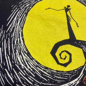 May include: Black t-shirt featuring a graphic from The Nightmare Before Christmas. The design includes a silhouette of Jack Skellington in front of a yellow full moon, with swirling white and black details. Orange pumpkins are visible at the bottom.