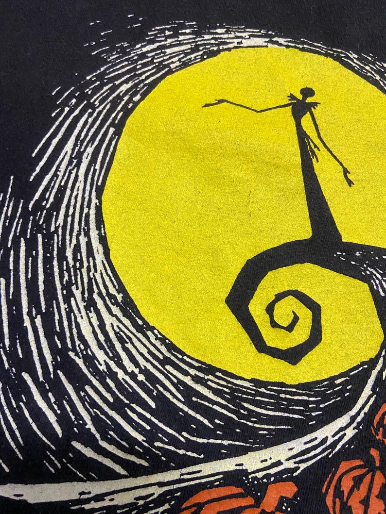 May include: Black t-shirt featuring a graphic from The Nightmare Before Christmas. The design includes a silhouette of Jack Skellington in front of a yellow full moon, with swirling white and black details. Orange pumpkins are visible at the bottom.