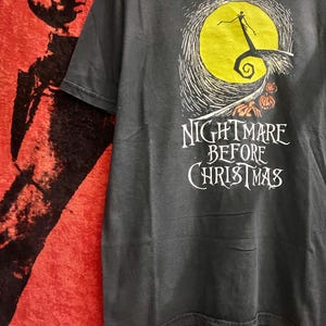 May include: Dark gray t-shirt featuring a graphic from "The Nightmare Before Christmas." The design includes a yellow full moon, a silhouetted figure, and the text "Nightmare Before Christmas" in white. The shirt is short-sleeved.