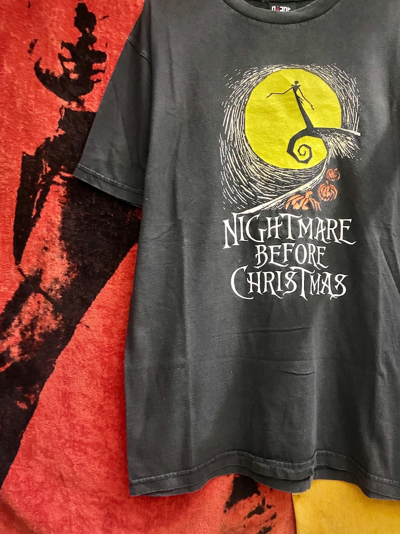 May include: Dark gray t-shirt featuring a graphic from "The Nightmare Before Christmas." The design includes a yellow full moon, a silhouetted figure, and the text "Nightmare Before Christmas" in white. The shirt is short-sleeved.