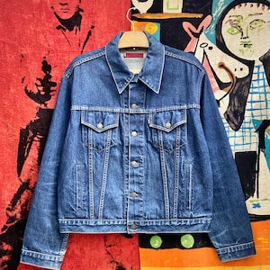 Könnte beinhalten: Eine klassische blaue Jeansjacke mit Knopfleiste und zwei Brusttaschen. Die Jacke hängt an einem Holzbügel. Der Denim hat eine mittlere Waschung mit dezenten Ausbleichungen. Der Kragen ist ein klassischer Spitzkragen.