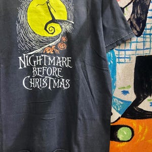 May include: Dark gray t-shirt featuring a graphic from "The Nightmare Before Christmas." The design includes a yellow moon, Jack Skellington, and the film's title in white lettering. The shirt is a short-sleeved crew neck.