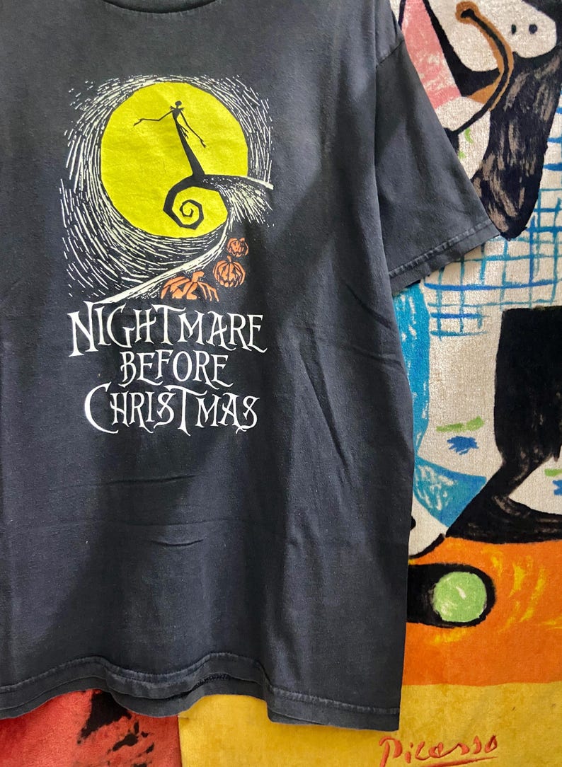 May include: Dark gray t-shirt featuring a graphic from "The Nightmare Before Christmas." The design includes a yellow moon, Jack Skellington, and the film's title in white lettering. The shirt is a short-sleeved crew neck.