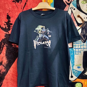 Könnte beinhalten: Schwarzes T-Shirt mit einem farbenfrohen Grafikdesign einer Kreatur, die eine Waffe hält, darunter das Wort "VIOLATOR" in stilisierten Buchstaben. Das T-Shirt hängt an einem Holzkleiderbügel.