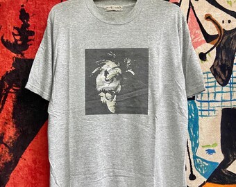 May include: A grey t-shirt with a black and white image of a person's face on the front. The image is in a square frame. The t-shirt has a crew neck and short sleeves.