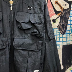Peut inclure: Veste de bombardier noire avec plusieurs poches avant et une fermeture &eacute;clair argent&eacute;e. La veste pr&eacute;sente un col et des poignets c&ocirc;tel&eacute;s, et un petit &eacute;cusson logo sur le bas de la face avant. Le logo The North Face est visible.