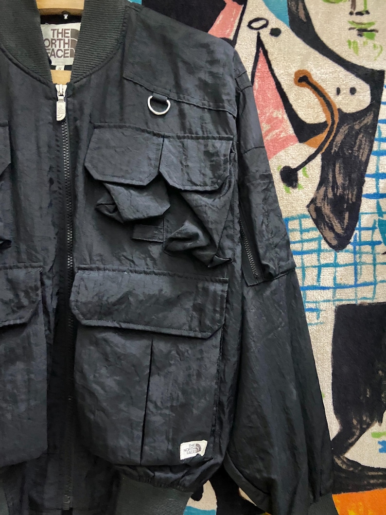 Peut inclure: Veste de bombardier noire avec plusieurs poches avant et une fermeture &eacute;clair argent&eacute;e. La veste pr&eacute;sente un col et des poignets c&ocirc;tel&eacute;s, et un petit &eacute;cusson logo sur le bas de la face avant. Le logo The North Face est visible.
