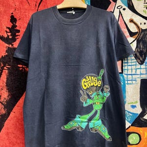 Könnte beinhalten: Dunkelblaues Marine-T-Shirt mit einer Cartoonfigur in grüner und blauer Kleidung und dem Wort "G&Us" in einer gelben Blase. Die Figur zeigt das Friedenszeichen. Das T-Shirt hängt an einem Holzbügel.