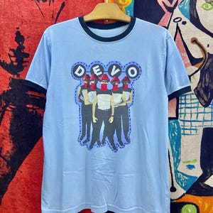 May include: Light blue ringer t-shirt featuring a graphic of the band DEVO. The graphic shows five figures with red hats and face coverings. The shirt has a black collar and sleeve trim, and is hanging on a wooden hanger.