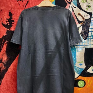 May include: A dark gray t-shirt hanging on a wooden hanger. The short-sleeved shirt has a crew neck and appears to be made of a soft, worn material. The background includes colorful artwork and a red rug.