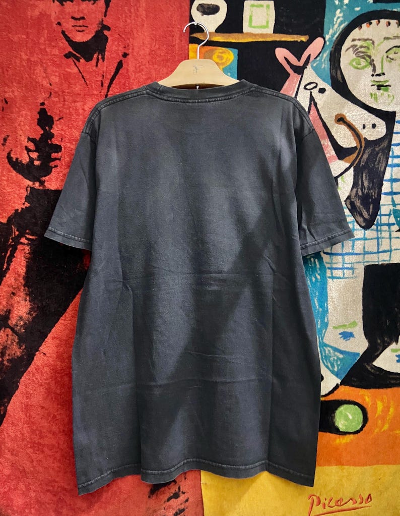 May include: A dark gray t-shirt hanging on a wooden hanger. The short-sleeved shirt has a crew neck and appears to be made of a soft, worn material. The background includes colorful artwork and a red rug.