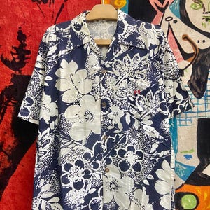 Puede incluir: Una camisa de manga corta con fondo azul marino y grandes estampados florales blancos. La camisa tiene un cuello clásico y botones de madera. Un pequeño acento rojo es visible en el bolsillo del pecho.