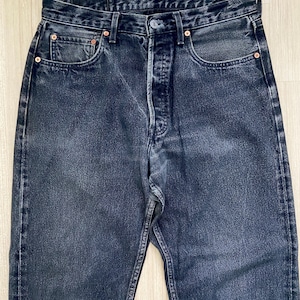 May include: A close-up shot of dark-wash denim jeans. The jeans feature a button fly, five-pocket design, and copper rivets. The denim has a faded, worn look, suggesting a vintage or pre-owned item. A classic style of jeans.