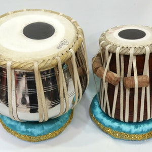 May include: A pair of traditional Indian tabla drums, one with a silver finish and the other with a brown wooden finish. Both drums are decorated with white leather straps and have a blue cushion underneath.