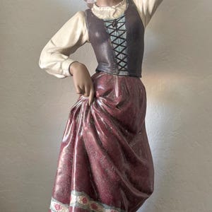 May include: A ceramic figurine of a person wearing a headscarf, a cream-colored long-sleeved blouse, a dark brown lace-up bodice, and a long maroon skirt with a floral border, balancing a brown jug on their head while standing on a green base.