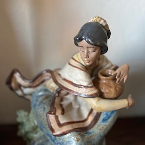 May include: A ceramic figurine of a woman in a flowing blue dress and cream-colored shawl, holding a brown pot. The figurine has dark hair and is in a seated pose, with a serene expression. The dress has a blue floral pattern.