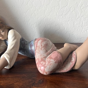 May include: A porcelain figurine of a reclining figure with brown hair, wearing a white long-sleeved top and pink patterned trousers. The figure rests on one arm with the other arm extended forward, while the legs are bent at the knees.