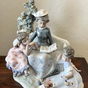 May include: A porcelain figurine depicting a woman reading a book to two children and a dog. The woman is wearing a grey dress and a hat. The children are wearing dresses with polka dots and floral patterns. The dog is brown and white.