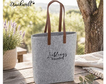 Könnte beinhalten: Eine graue Filz-Tragetasche mit braunen Ledergriffen. Auf der Tasche steht in schwarzer Schrift "Lieblingsfreundin". Die Tasche steht auf einem Holztisch mit einer Pflanze in einem geflochtenen Topf.