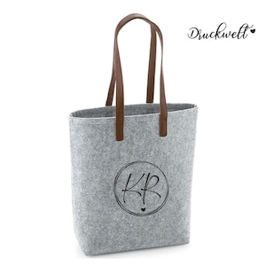 Könnte beinhalten: Graue Filz-Tragetasche mit braunen Ledergriffen. Die Tasche hat ein kreisförmiges Design mit den Initialen "KR" in Schwarz und dem Wort "Druckwelt" in Schrift. Die Tasche ist zum Tragen von Gegenständen konzipiert.