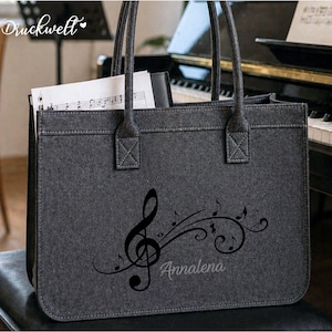 Könnte beinhalten: Graue Filz-Tragetasche mit schwarzen Noten und einem Notenschlüssel-Design. Die Tasche hat zwei Griffe und enthält Noten. Der Name "Annalena" ist auf der Tasche aufgedruckt.