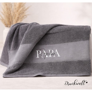Könnte beinhalten: Ein graues Badetuch mit einem helleren grauen Streifen in der Mitte. Das Wort "PAPA" und der Name "Sophia" sind in weißer Schrift auf den Streifen gestickt. Das Handtuch ist gefaltet, und in der unteren rechten Ecke steht "Druckwelt".