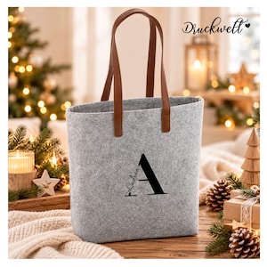 May include: A light grey felt tote bag with two brown faux leather handles. The front of the bag features a large black letter A with the name Andrea written in a cursive script across the letter. The text Druckwelt is visible.