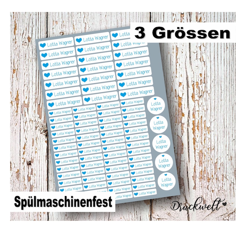 May include: A sheet of grey adhesive labels featuring the name Lotta Wagner in blue text alongside a blue heart icon. The sheet includes rectangular and circular labels in three sizes, with the text 3 Gr&ouml;ssen and Sp&uuml;lmaschinenfest visible.