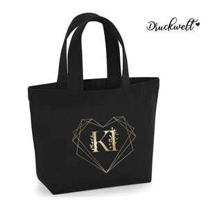 May include: Black canvas tote bag with gold foil lettering and a geometric heart design. The letters "KI" are inside the heart.
