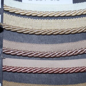 May include: A sample card displaying five different colours of trim, labelled "Ecru", "H. Yellow", "Beige", "Camel", and "W. Gold". The trim is a combination of a woven fabric and a braided cord.