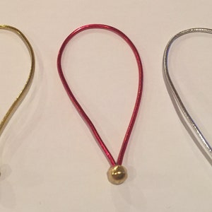 Peut inclure: Trois boucles de cordon élastique avec des perles métalliques dorées aux extrémités. Les cordons sont dorés, rouges et argentés.
