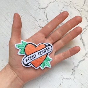 Puede incluir: Un corazón naranja con una pancarta blanca que dice "AGENT COOPER" rodeado de hojas verdes. El corazón es un parche.