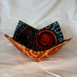 May include: A handmade fabric bowl with a unique design. The bowl features a vibrant mix of colors and patterns, including orange, red, blue, and black. The bowl is designed to hold hot items, such as soup or oatmeal.