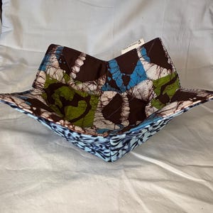 May include: A square, fabric bowl with a colorful batik pattern. The bowl features a brown, blue, green, and white design. The interior is a mix of brown, blue, and green. The exterior has a blue and white pattern.