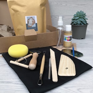 May include: A pottery kit containing wooden tools, a sponge, and metallic acrylic paint. Also included are a spray bottle, a paintbrush, and a succulent plant in a grey pot. The tools are arranged on a black cloth. The kit is presented in a cardboard box.