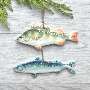 May include: Two ceramic fish ornaments. The top fish is green, white, and orange, while the bottom fish is blue and white. Both are suspended by twine and set against a white wood background with a sprig of green foliage.