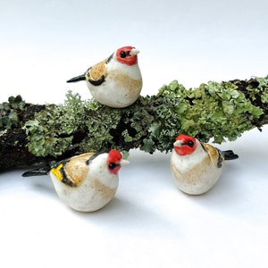 May include: Three ceramic bird figurines perched on a branch covered in green moss. The birds are painted with white, black, and yellow accents and have red heads.