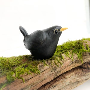 May include: A handmade blackbird ornament with a yellow beak and detailed eye. The bird is positioned on a piece of driftwood covered in green moss. The bird is approximately 7.6 cm long, creating a naturalistic scene.