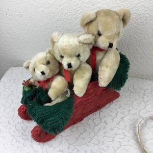 Puede incluir: Tres osos de peluche beige de diferentes tamaños sentados dentro de un trineo decorativo rojo. Cada oso lleva una cinta roja fina alrededor del cuello. El trineo está decorado con tela de ganchillo verde oscuro y bayas rojas pequeñas.