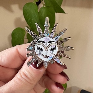 May include: A silver-coloured enamel pin in the shape of a sun with a face design. The pin features a white face with black details and iridescent accents. The sun has pointed rays extending outwards.