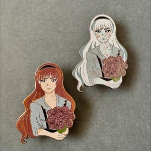 May include: Two enamel pins featuring illustrations of women holding bouquets of flowers. The women have long hair and are wearing striped shirts. The pins are in shades of red, white, and gray, with a touch of pink.